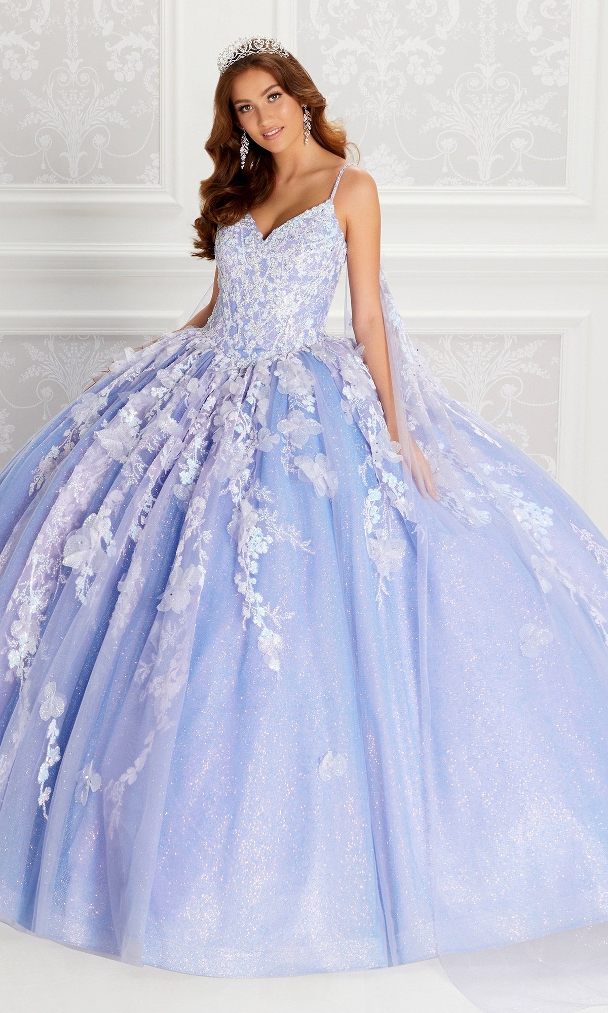 A woman in the Quinceanera Dress PR22143 By Princesa, featuring a sparkling lavender hue, beaded corset bodice, and floral appliqué details, with a tiara, stands before an ornate white background.